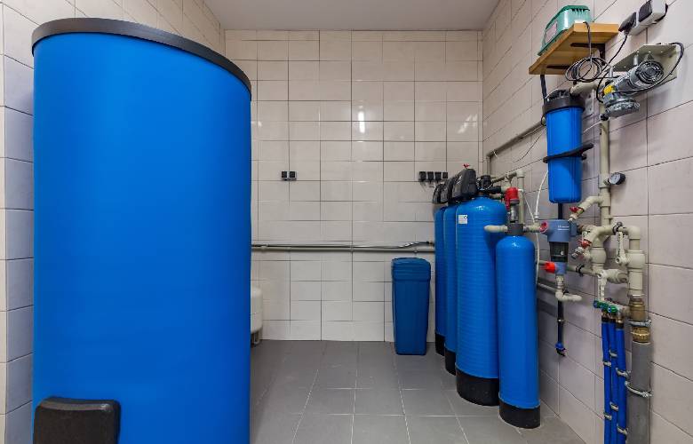 Een boilerruimte met een wit betegelde muur en een grijs betegelde vloer. In de ruimte staan verschillende blauwe Clearwater waterverzachters en andere systemen voor waterbehandeling.